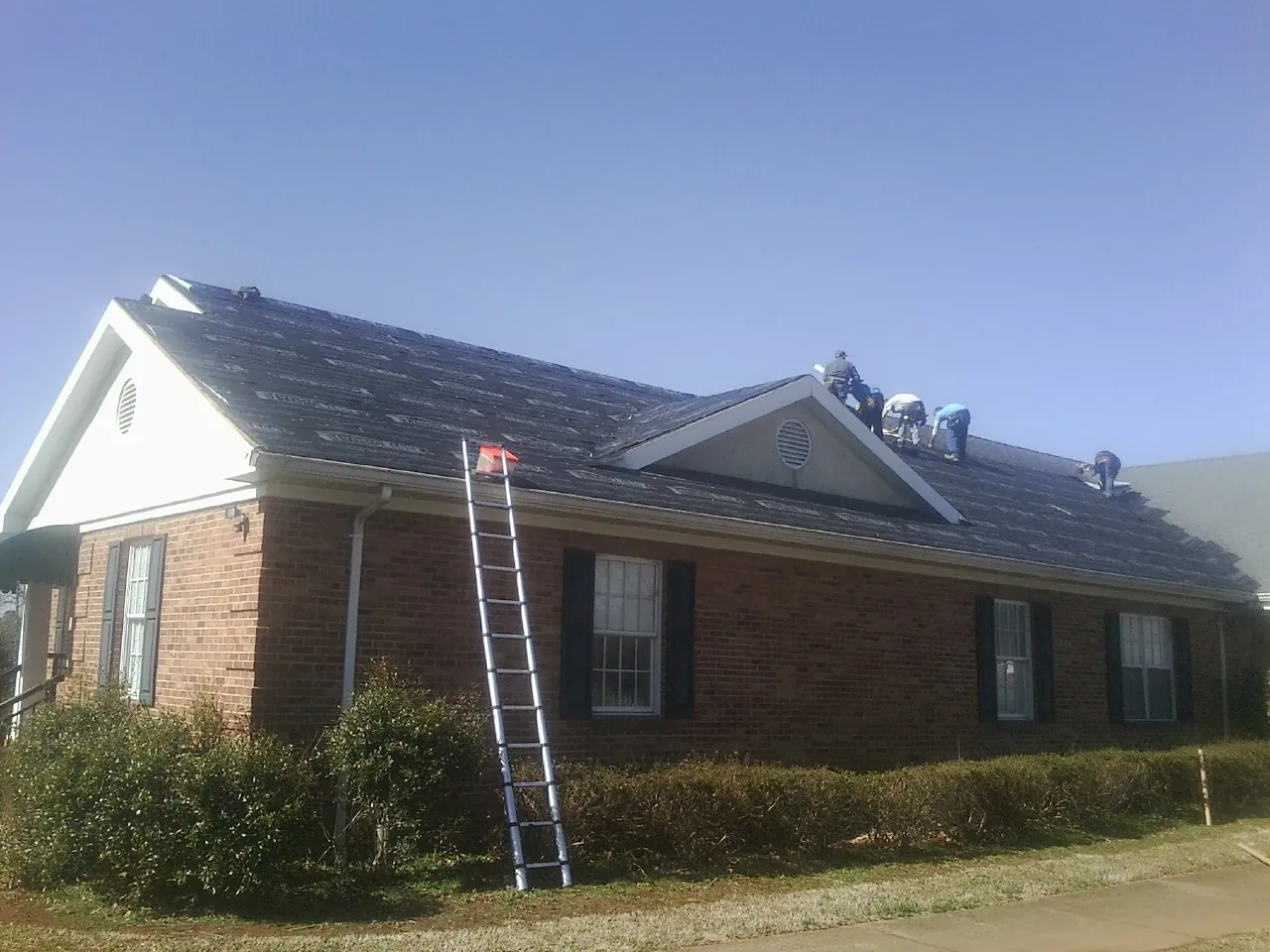 Crew tearing shingles off a brick ranch roof with a ladder set — Metal Roof Repair work by our crew in Mckenna