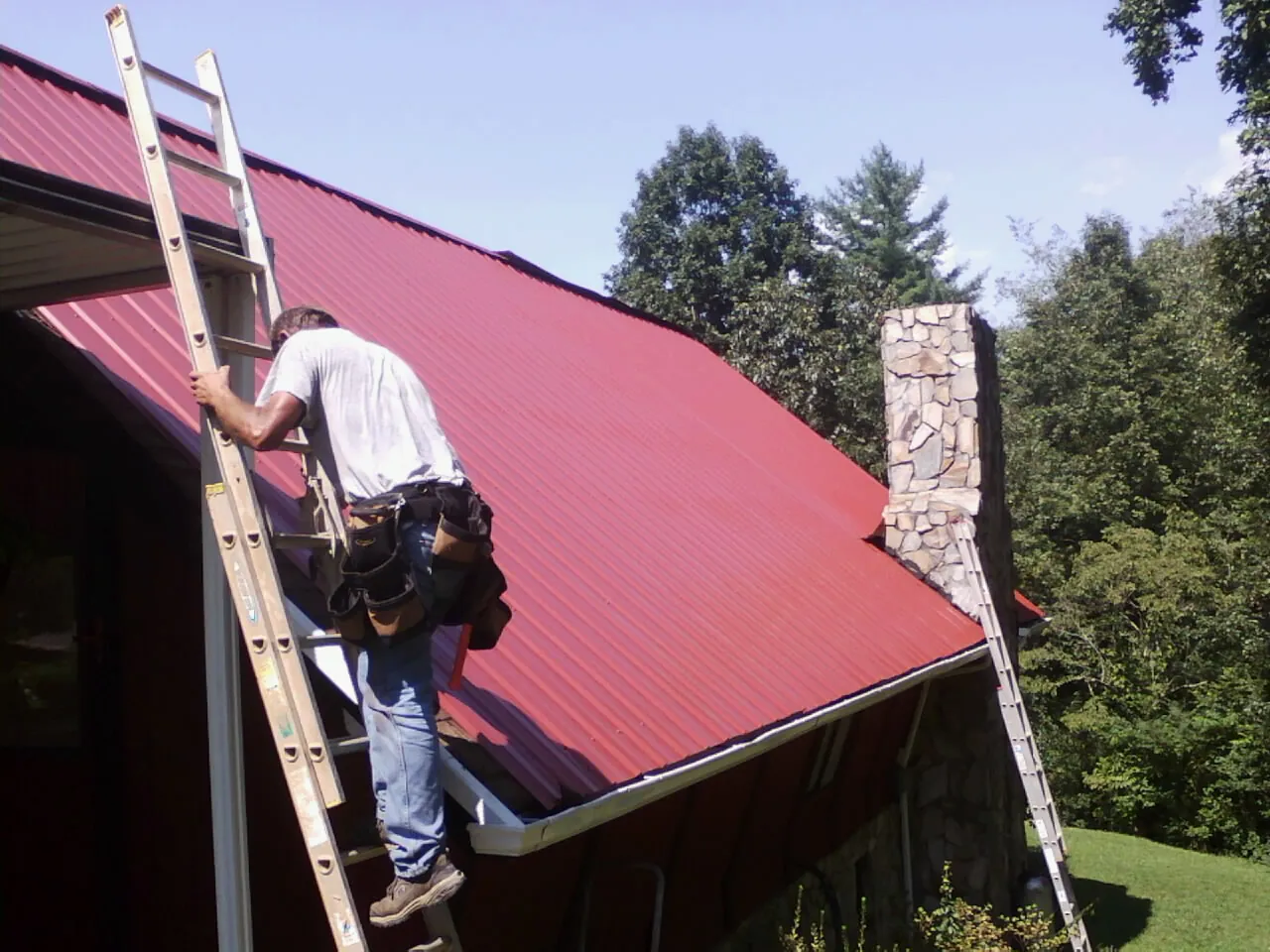 Roofer on a ladder beside a red metal roof and stone chimney — Soffit & Fascia Repair by Roofing Brothers in Mckenna